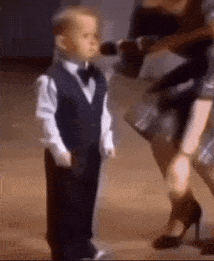 a little boy in a tuxedo and bow tie is standing next to a woman holding a microphone .