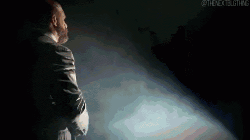 a man in a suit is standing in front of a spotlight in a dark room with smoke coming out of it .
