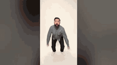 a man with a beard is jumping in the air while wearing a grey shirt and black pants .