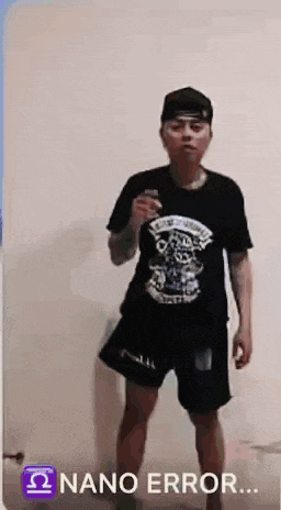 a man wearing a black shirt and black shorts is standing in front of a white wall holding a cell phone .