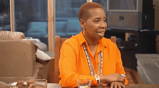 a woman in an orange shirt is sitting at a table with a glass of water and a watch .