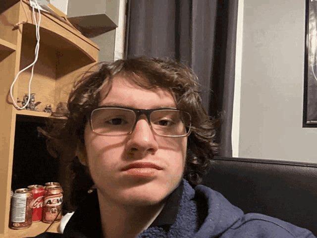 a young man wearing glasses takes a selfie in front of a shelf with cans of coca cola