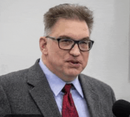 a man wearing glasses and a tie is speaking into a microphone .