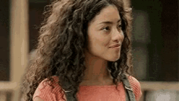 a young woman with curly hair is wearing overalls and a pink shirt and smiling .