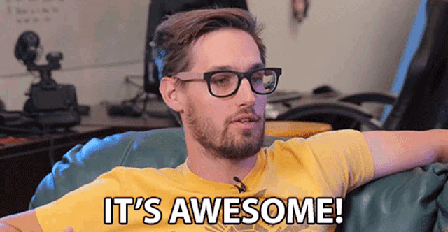 a man wearing glasses and a yellow shirt is sitting on a couch and says it 's awesome