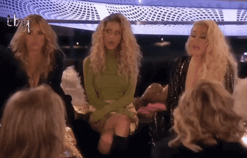 a group of blonde women are sitting in a circle with one wearing a green turtleneck