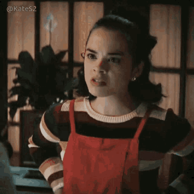 a girl wearing a red apron and a striped sweater is standing in front of a window