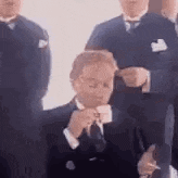 a man in a suit and tie is smoking a cigarette while standing in front of a group of men .