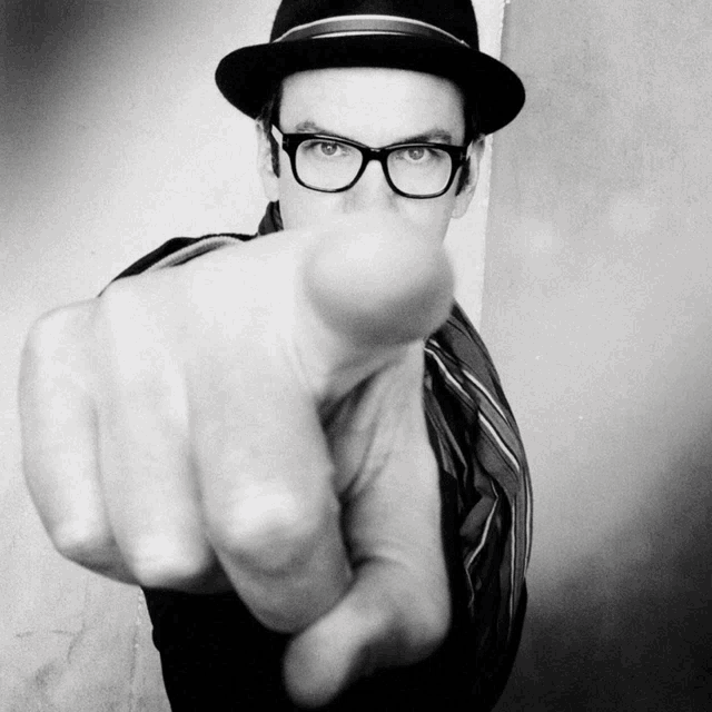 a man wearing glasses and a hat is pointing his finger at the camera