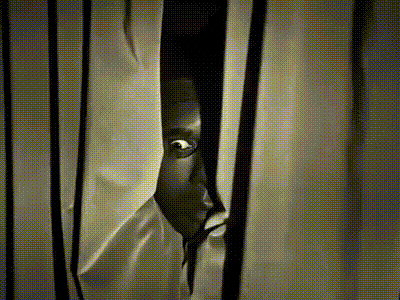 a black and white photo of a person peeking through a window curtain .