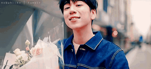 a young man in a denim jacket is holding a bouquet of flowers and smiling .
