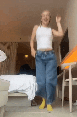 a woman in a white tank top and blue jeans is jumping in the air in a room .