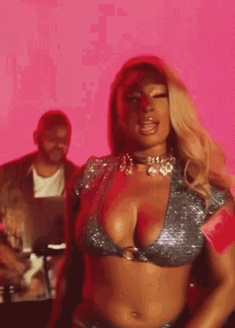 a woman in a bikini top is dancing in front of a pink background while a man looks on .