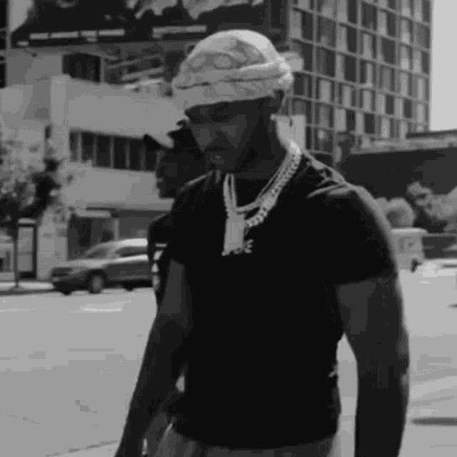 a black and white photo of a man wearing a bandana and a necklace walking down a street .