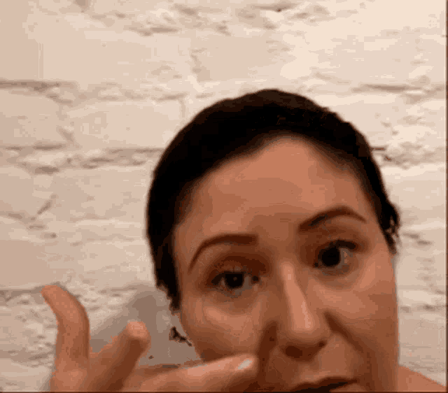 a woman wipes her face with her hand in front of a white brick wall