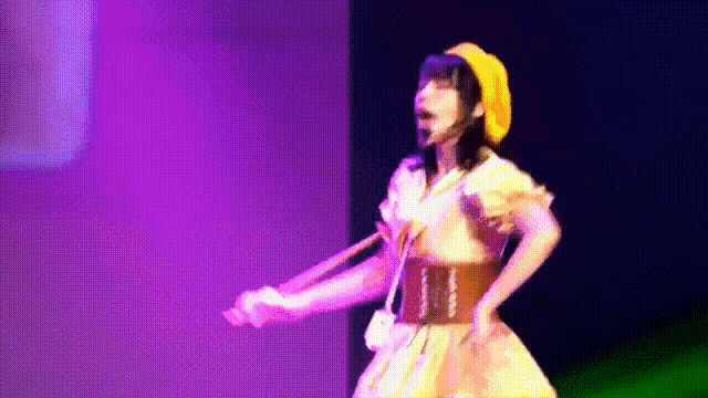 a woman in a yellow dress and yellow hat is dancing on a stage with purple lights behind her .