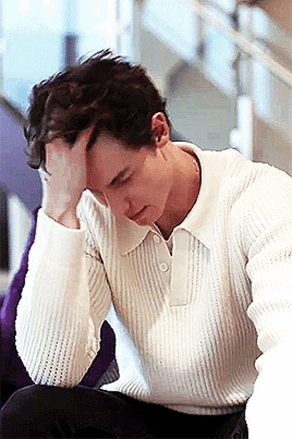 a young man in a white sweater is sitting down with his hand on his head .