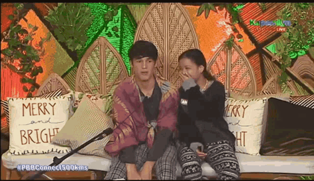 a boy and a girl sit on a couch with merry and bright pillows