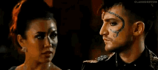 a man and a woman are looking at each other in a dark room . the man has a tattoo on his face .