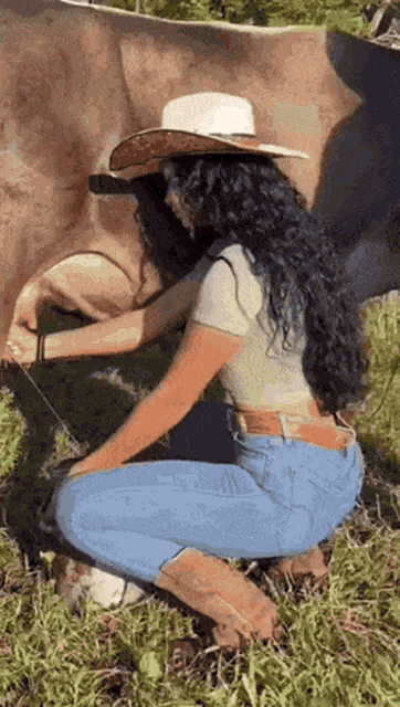 a woman in a cowboy hat is kneeling down next to a cow in a field .