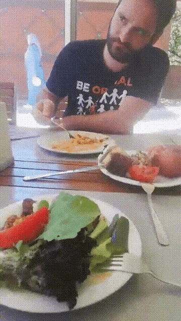 a man wearing a black shirt that says be or al sits at a table with plates of food