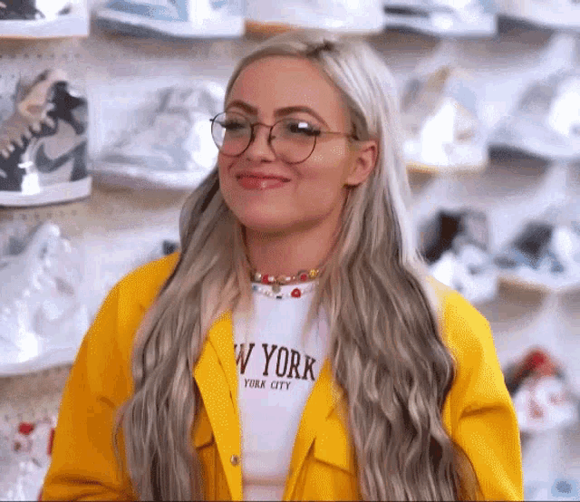 a blonde woman wearing glasses and a yellow jacket is standing in front of a wall of shoes .
