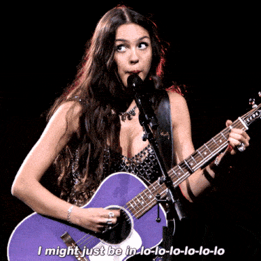 a woman singing into a microphone while holding a purple guitar that says i might just be in