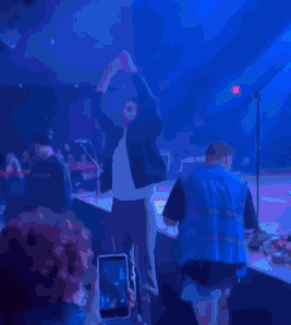 a man is standing on a stage with his hands behind his head while a man in a vest stands behind him .