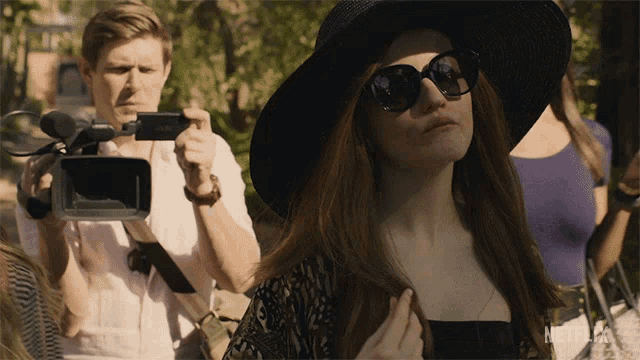 a woman wearing sunglasses and a hat stands in front of a man holding a camera and a sign that says netflix