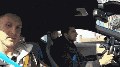 two men are driving a car and one of them is wearing a blue belt .