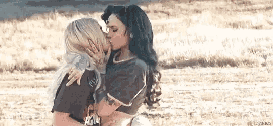two women are kissing in a field while standing next to each other .