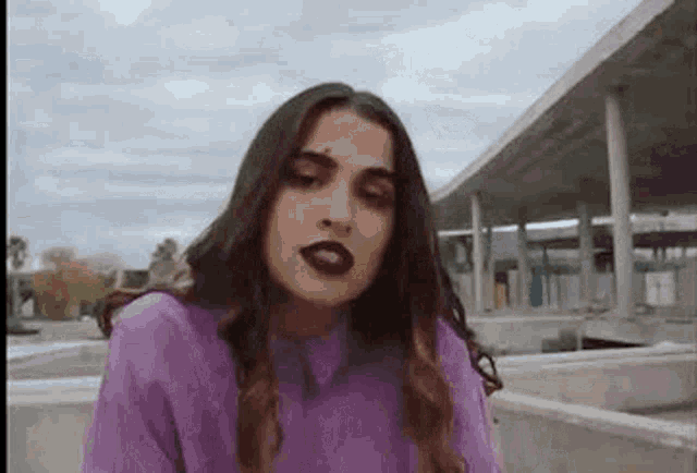 a woman in a purple shirt and dark lipstick is looking at the camera .