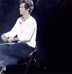 a man in a white shirt and black pants is sitting on a stage holding a microphone .