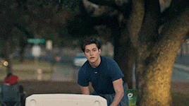 a young man in a blue shirt is sitting in front of a tree holding a cooler
