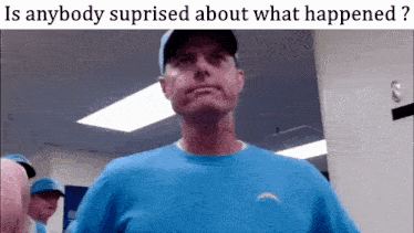 a man in a blue shirt is standing in front of a wall with the words is anybody suprised about what happened