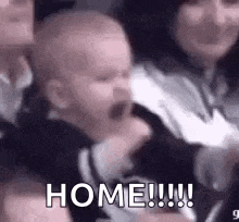 a baby is yawning while sitting in a crowd of people and a woman is holding it .