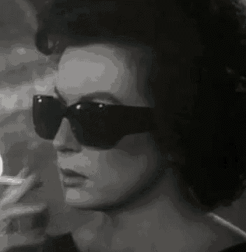 a black and white photo of a woman wearing sunglasses smoking a cigarette .