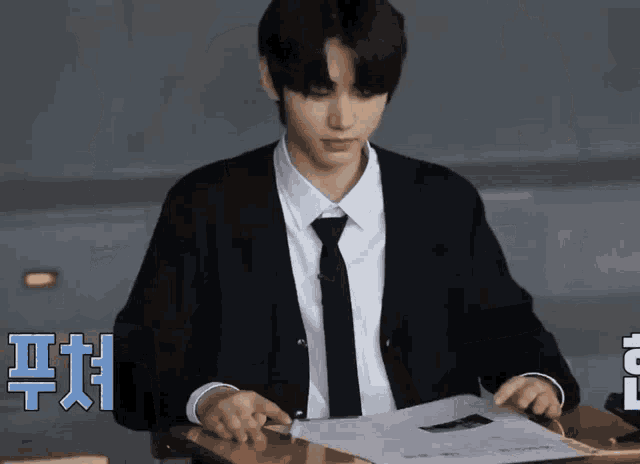 a man in a suit and tie is sitting at a desk in a classroom looking at a piece of paper .