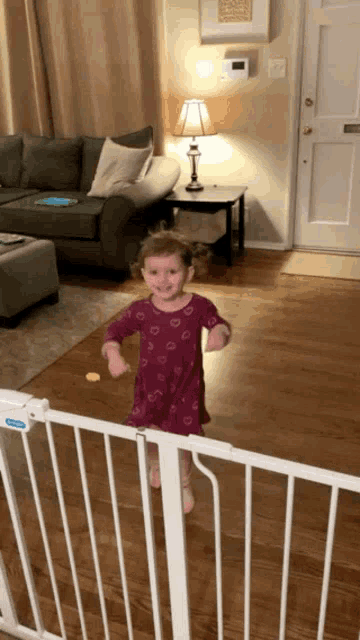 a little girl in a purple dress is standing in a living room behind a white gate