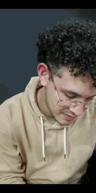 a young man with curly hair wearing glasses and a tan sweatshirt