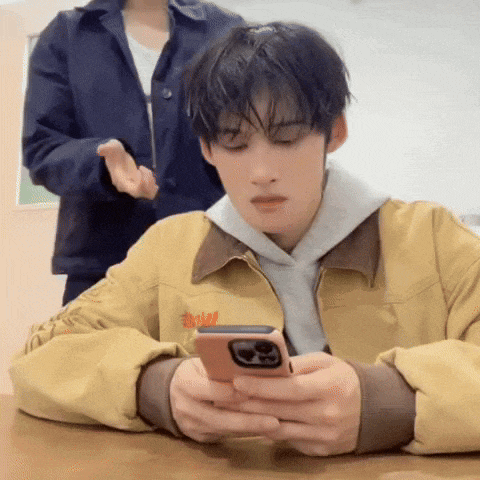 a man in a yellow jacket is sitting at a table looking at his cell phone .