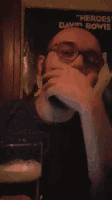 a man with glasses holds a glass of beer in front of a poster for david bowie