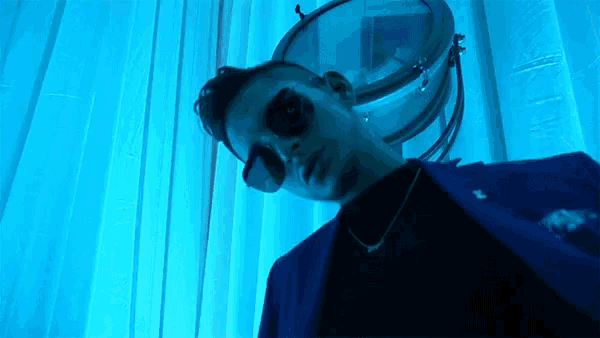 a man wearing sunglasses and a necklace is standing in front of a blue light .