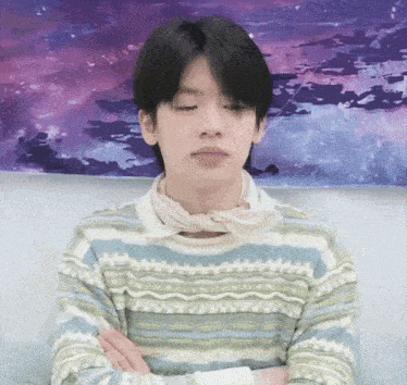 a young man wearing a striped sweater and a scarf around his neck is sitting with his arms crossed .