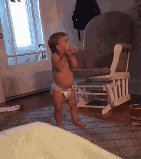 a baby in a diaper is standing next to a rocking chair
