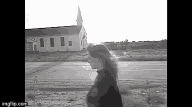a black and white photo of a woman walking down a road in front of a church .