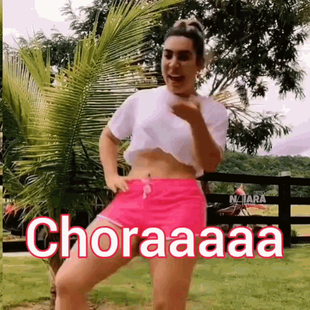 a woman in a white crop top and pink shorts is dancing in front of a palm tree .