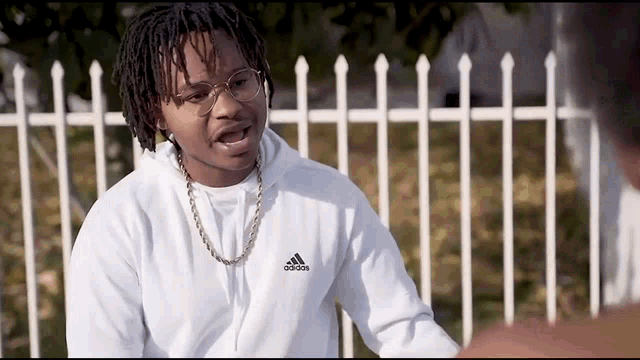 a man wearing a white adidas hoodie stands in front of a white fence