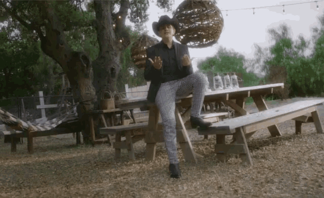a man in a cowboy hat is sitting on a wooden picnic table with his legs crossed .