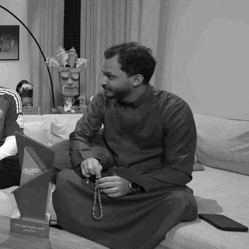 a black and white photo of a man sitting on a couch holding a rosary and a plaque with arabic writing